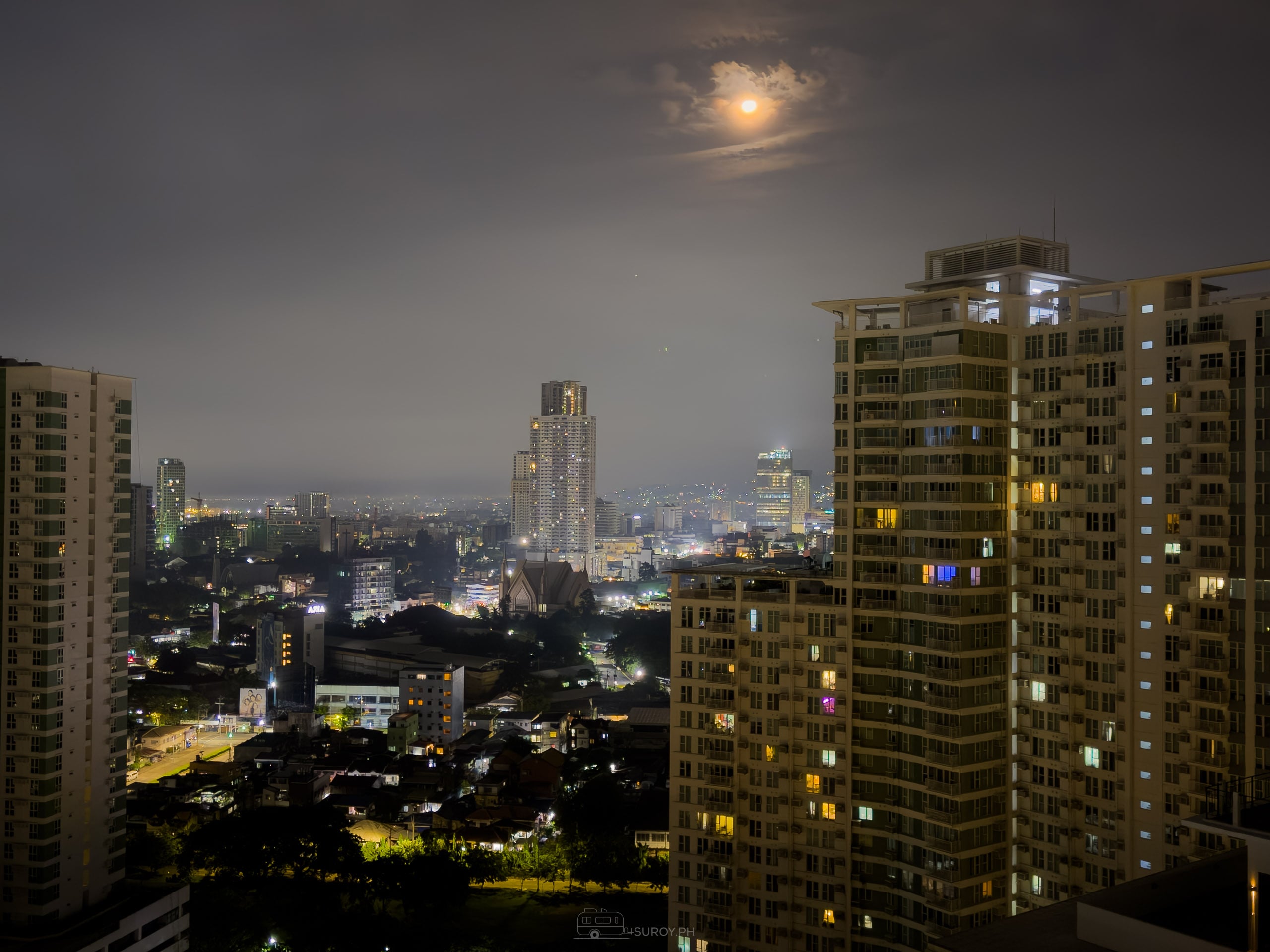 Cebu City at night on 21 September 2026, seen from a high floor. Lit condominium towers stand in the foreground, the moon shows a broad halo through the smoke layer, and the mid distance skyline and hills fade into flat gray.
