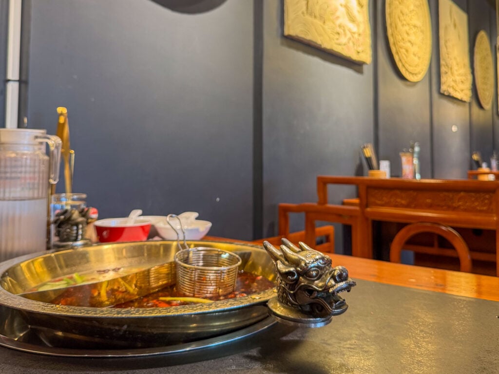 A brass divided hotpot with a cast dragon head on the rim sitting on a table at Long Men Hotpot Cebu, with woven round wall panels and wooden chairs in the empty dining room behind
