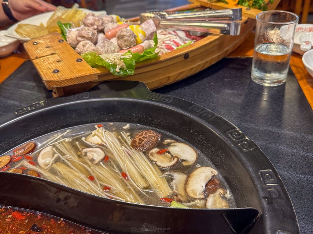 A wooden boat shaped platter of meatballs, sliced meats and vegetables on a table at Long Men Hotpot Cebu, above a divided pot of clear broth with mushrooms, corn and enoki