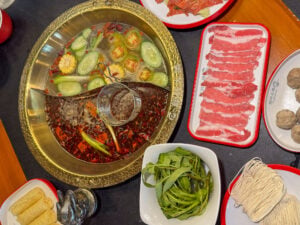 Overhead view of a divided brass hotpot at Long Men Hotpot Cebu, clear broth with cucumber and corn on one side and red chili broth on the other, surrounded by trays of sliced beef, meatballs, green beans, tofu and noodle nests