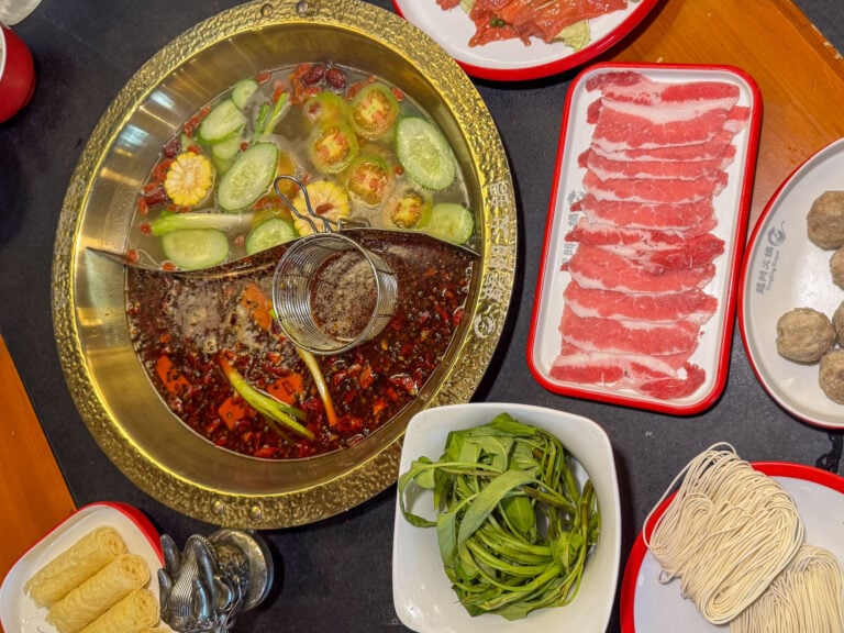 Overhead view of a divided brass hotpot at Long Men Hotpot Cebu, clear broth with cucumber and corn on one side and red chili broth on the other, surrounded by trays of sliced beef, meatballs, green beans, tofu and noodle nests