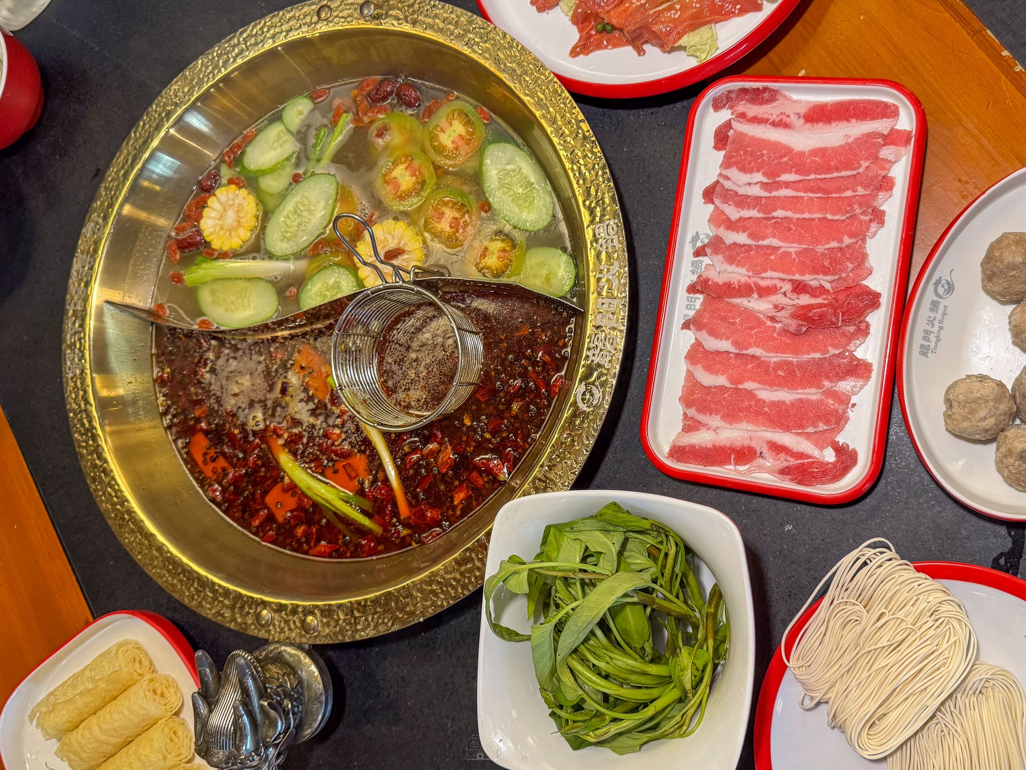 Overhead view of a divided brass hotpot at Long Men Hotpot Cebu, clear broth with cucumber and corn on one side and red chili broth on the other, surrounded by trays of sliced beef, meatballs, green beans, tofu and noodle nests