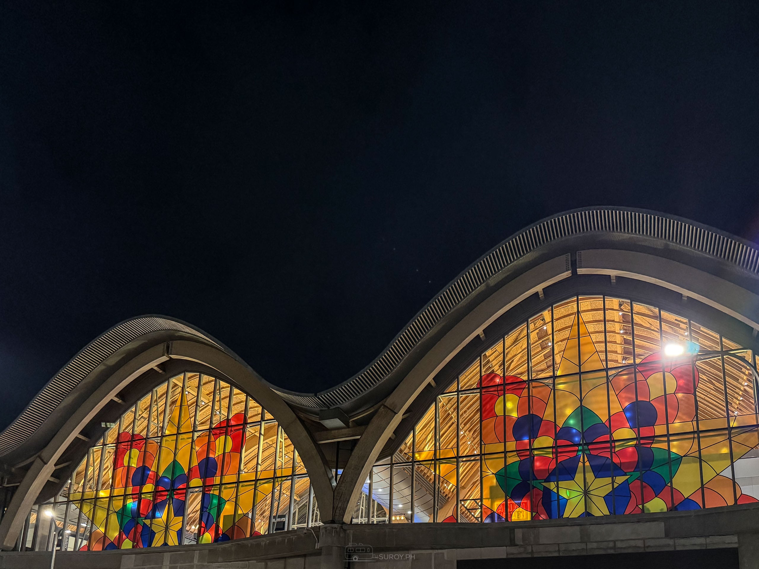 Two arched glass ends of Mactan-Cebu International Airport Terminal 2 side by side at night under the wave-shaped roof, both lit gold with a large colored star pattern