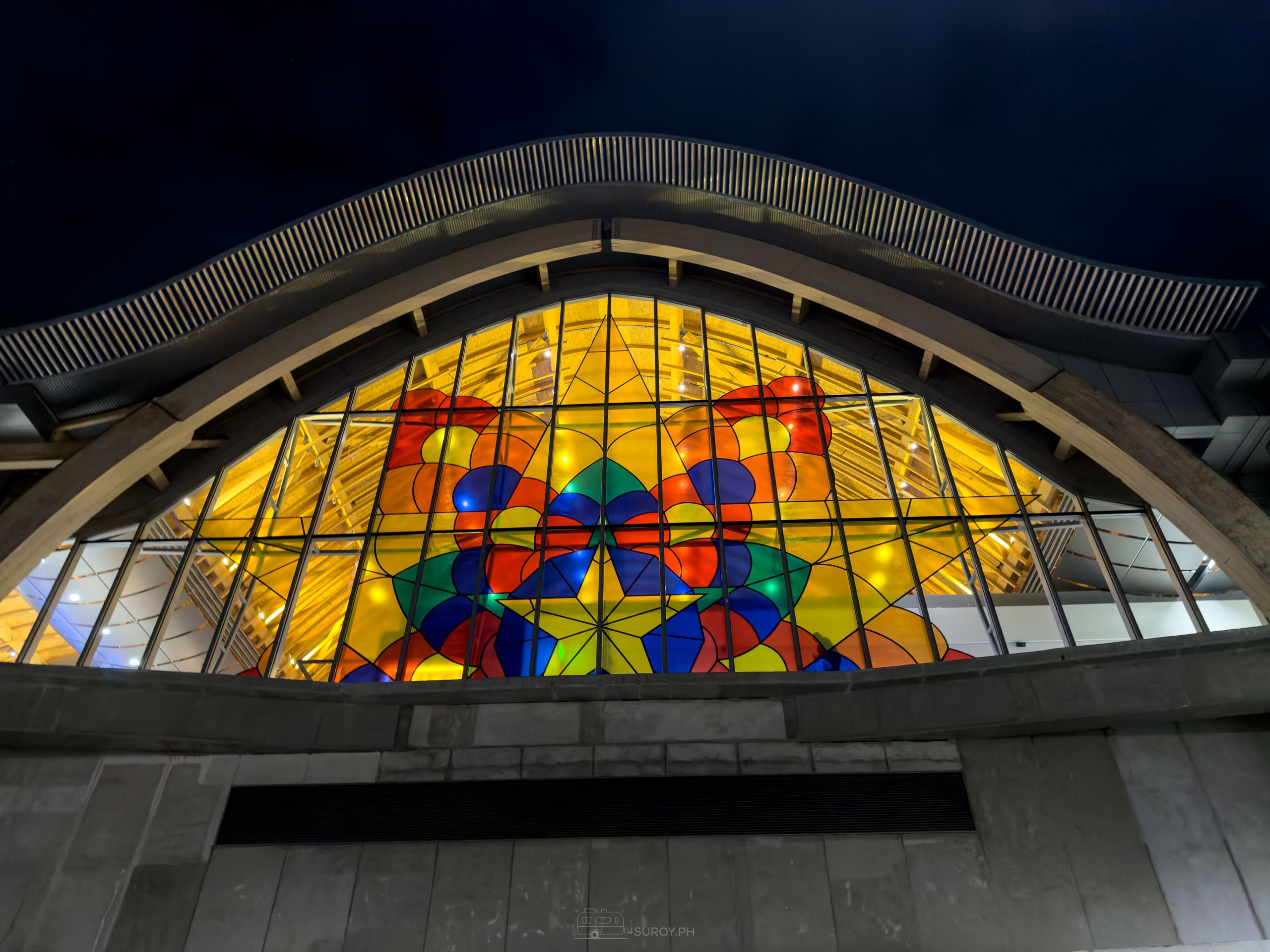 One arched glass end of Mactan-Cebu International Airport Terminal 2 at night, lit gold from inside with a large star pattern in red, blue, green and yellow, under the curved timber roof