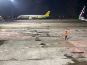 A marshaller in a reflective vest holds up red light wands on the apron at night, a Cebu Pacific jet parked under floodlights behind him and an aircraft winglet in the right corner