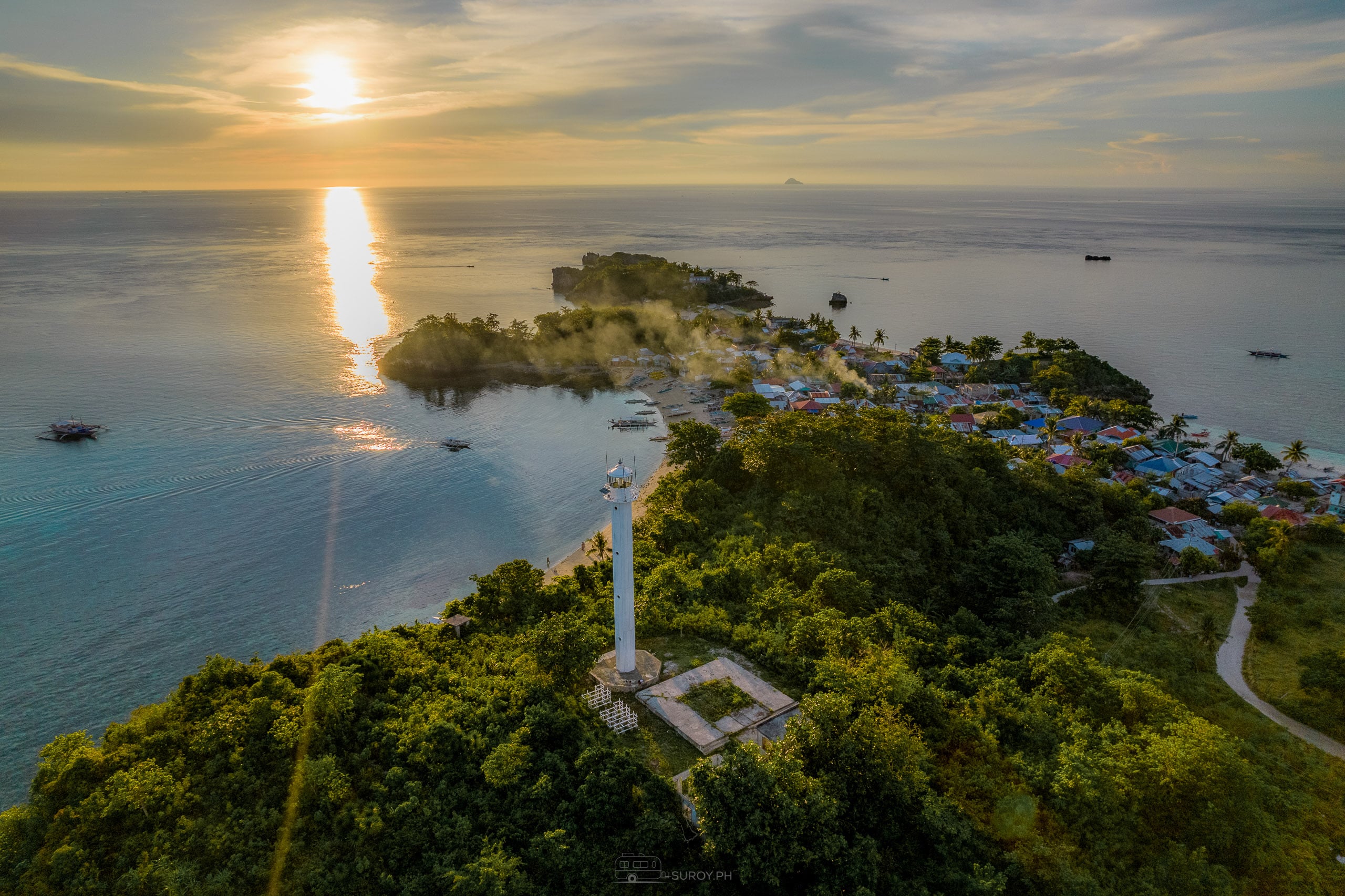 Aerial view over Malapascua Island at sunset, a white lighthouse on the wooded hill in the foreground, village roofs along the shore to the right, and the sun path running across the water past a small wooded islet