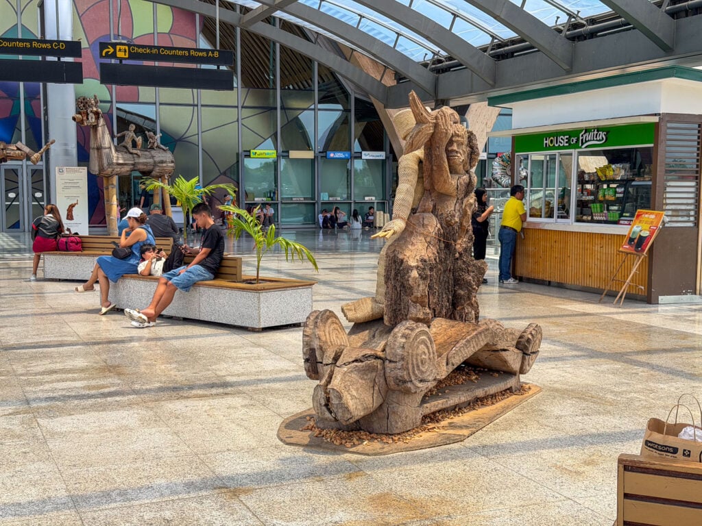 Travelers sitting on a stone bench beside a potted palm in Terminal 2 at Mactan Cebu International Airport, a tall carved wooden figure in the foreground and food kiosks behind.