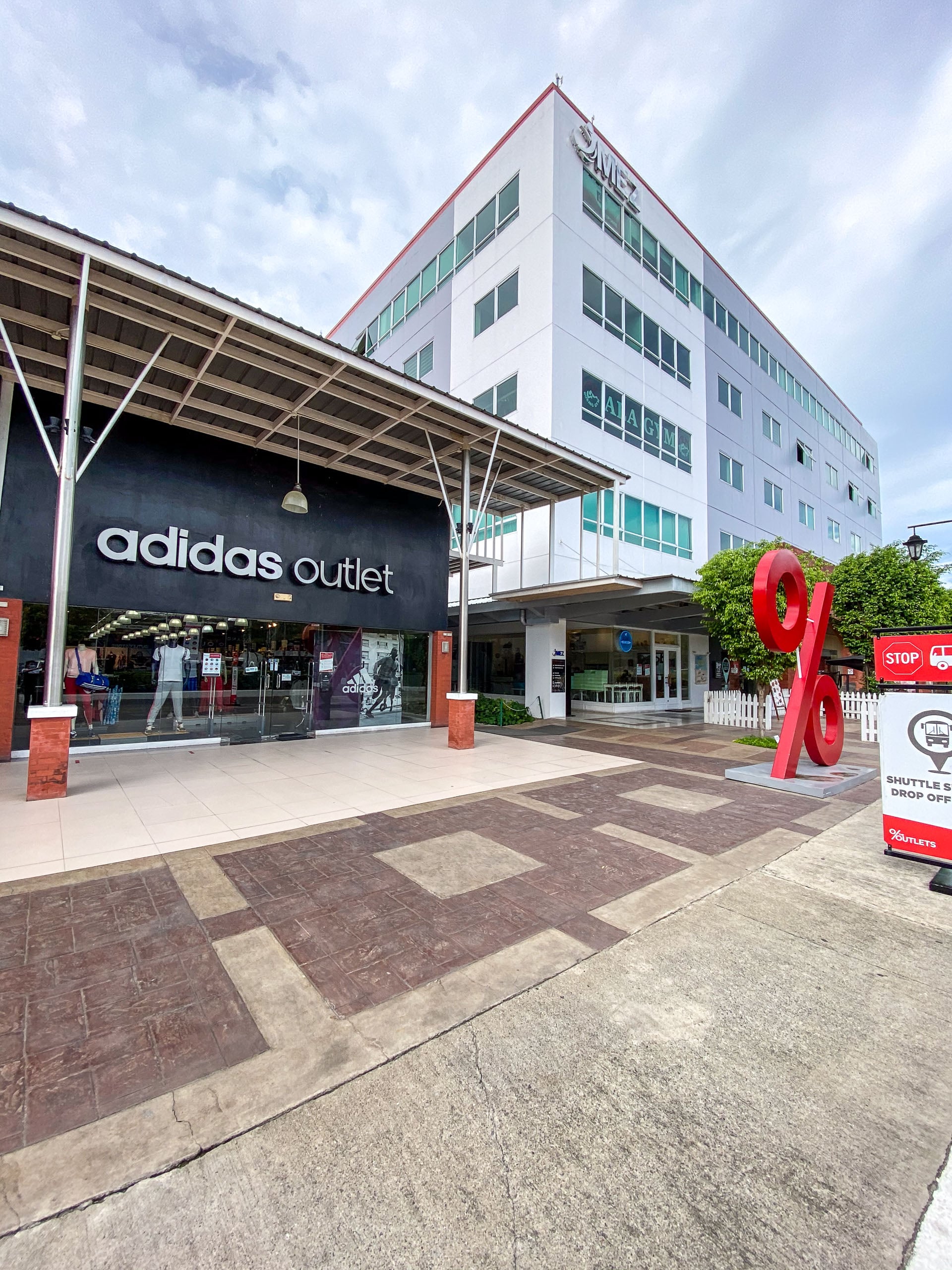 The Adidas Outlet storefront under a steel canopy, with a red percent-sign sculpture on the walkway and an office building behind