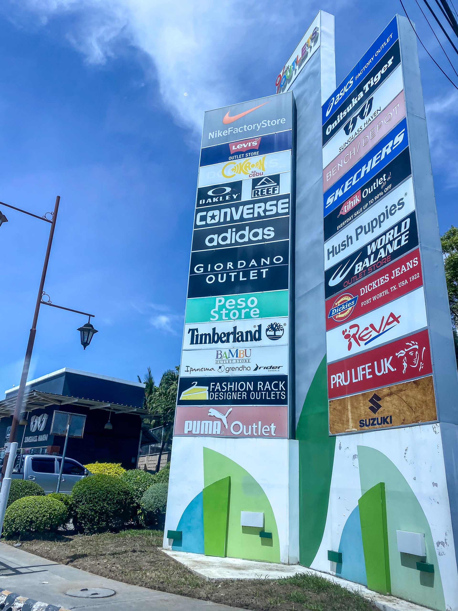 A tall roadside directory pylon listing outlet brands including Nike, Levis, Converse, Adidas, Timberland, Skechers and Hush Puppies, under a blue sky