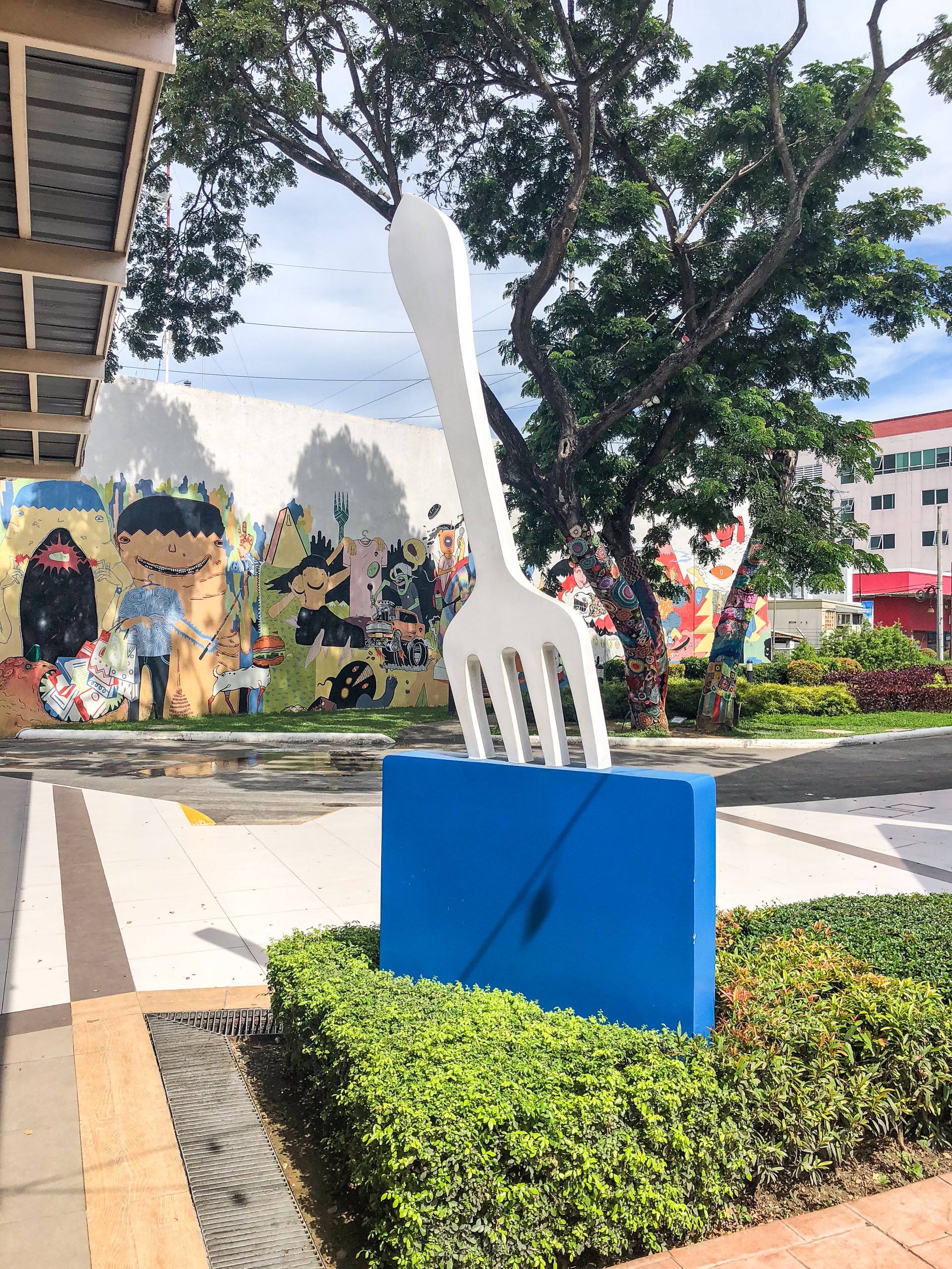A giant white fork sculpture on a blue base in a hedge, in front of a painted mural wall