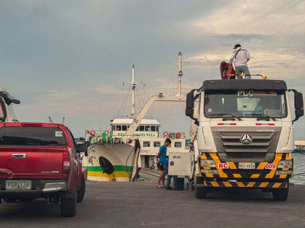A dump truck and a red pickup waiting on the pier to board a roll-on roll-off ferry at first light, with a man standing on the truck cab