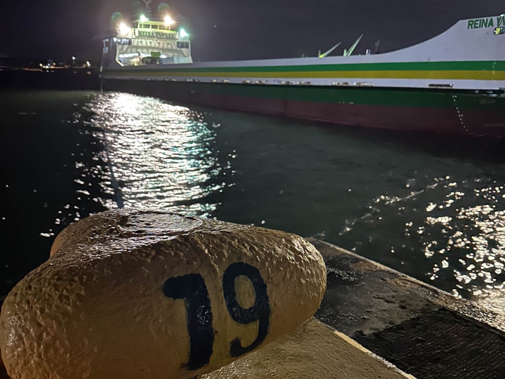 A mooring bollard numbered 19 in the foreground with a green and white ferry lying alongside behind it, deck lights reflecting on the water