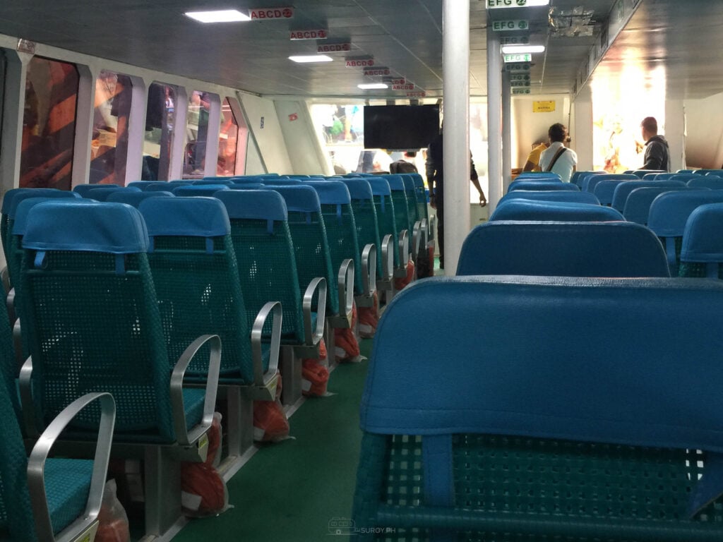 Rows of blue seats inside a fast craft passenger cabin with life vests stowed underneath and a few passengers seated toward the front