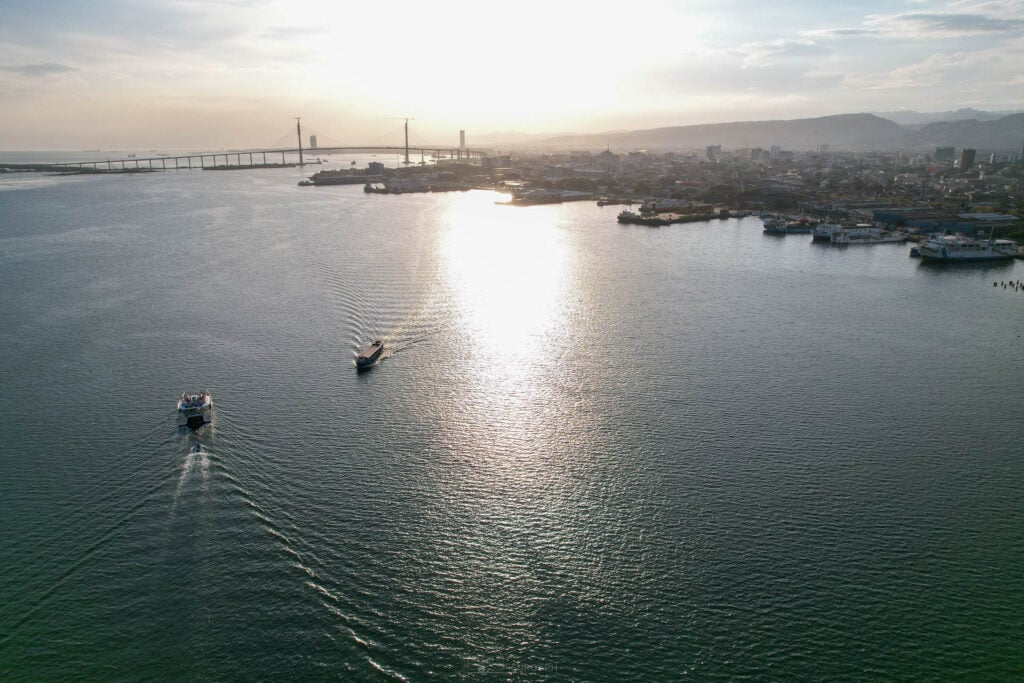 An aerial view of the Cebu channel at sunrise with a small boat drawing a wake across the light and the bridge on the horizon