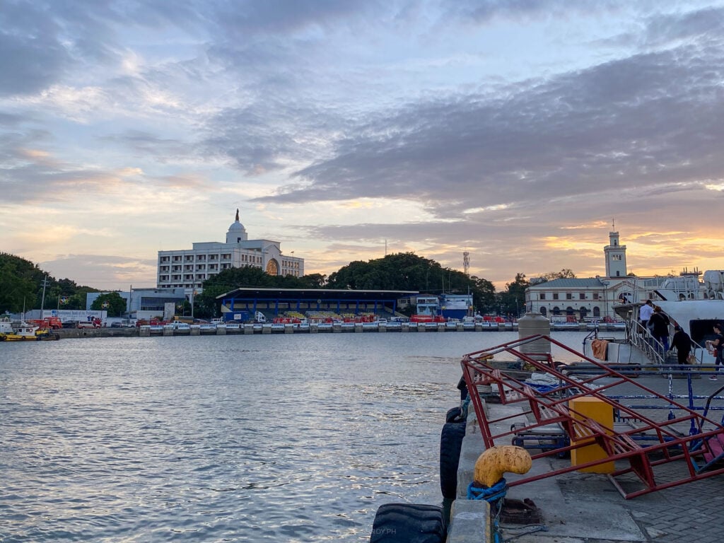 The lit Iloilo waterfront at dusk seen from a ferry, with the red steel of the ramp in the foreground