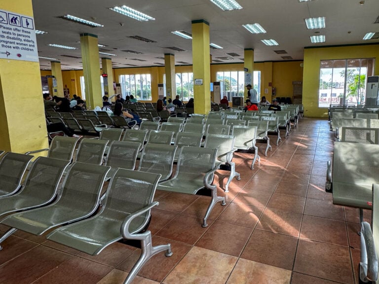 Rows of empty green metal seats in a ferry terminal waiting hall at Iloilo, a few passengers seated at the far end near the windows.