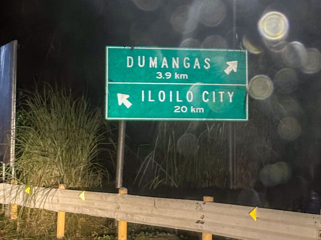 A roadside sign reading Dumangas 3.9 kilometres and Iloilo City 20 kilometres, seen at night through a wet window