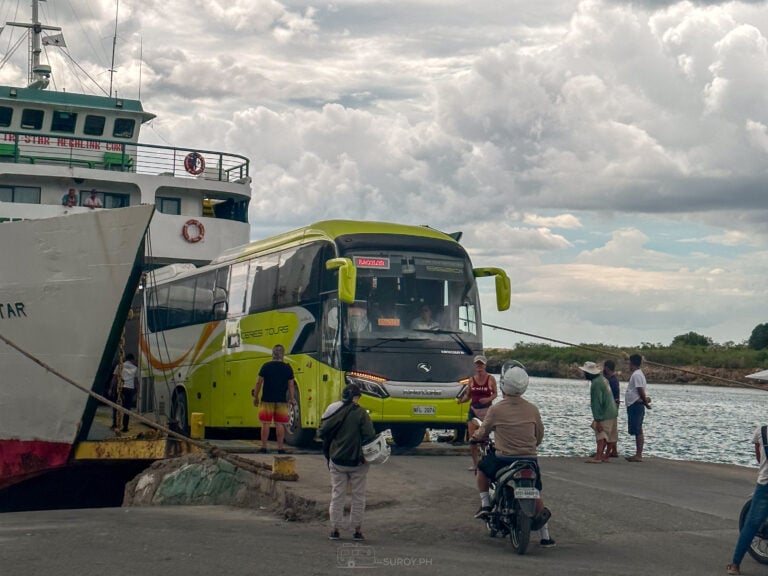 A tour bus reverses up the ramp of a RoRo ferry at a Philippine pier while foot passengers wait beside it holding their bags