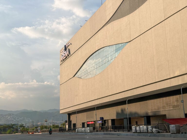 The beige curved facade of SM Seaside Cebu Arena with its Arena sign and a curved strip of glass, under a cloudy afternoon sky