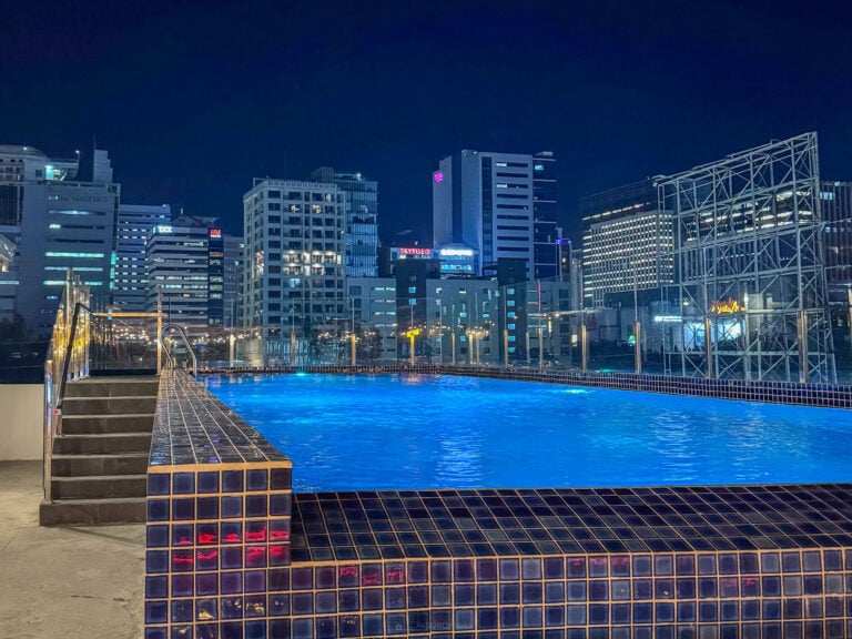 The rooftop pool at The Pad Co-Living in Cebu lit blue at night, with steps at one end and the lit towers and steel structures of the Banilad skyline behind
