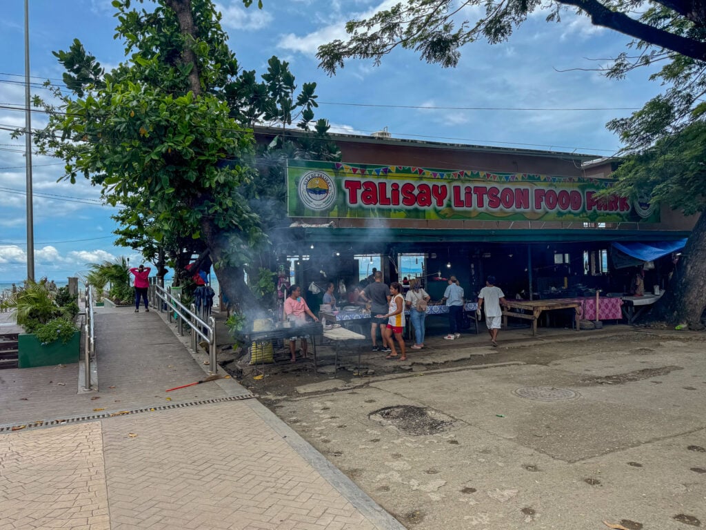 The paved approach to Talisay Litson Food Park with the signboard ahead and smoke rising from the stalls