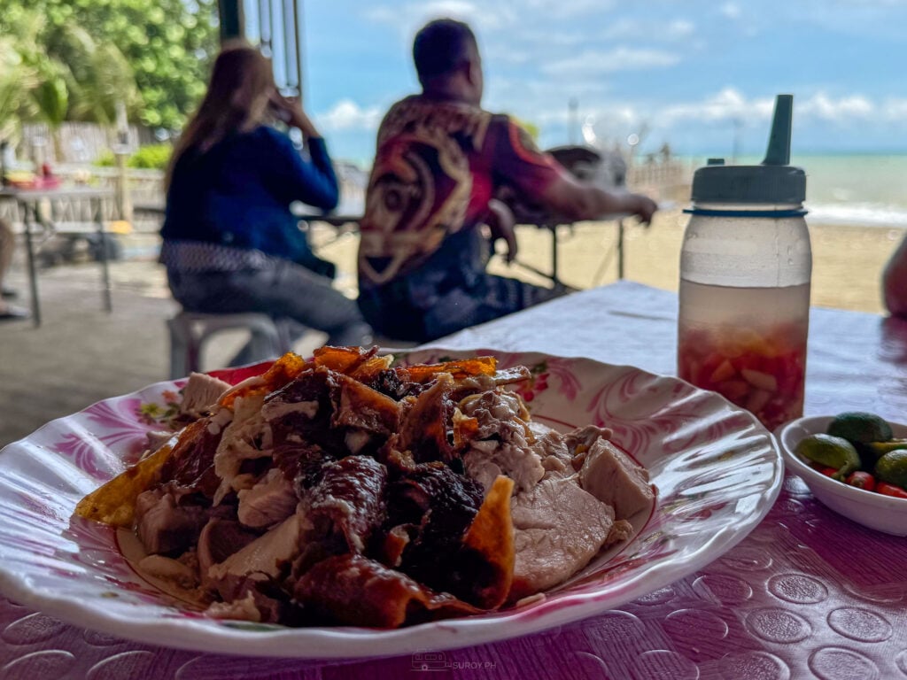 A plate of chopped Talisay lechon on a beachside table with a squeeze bottle of chilied vinegar and a dish of calamansi beside it
