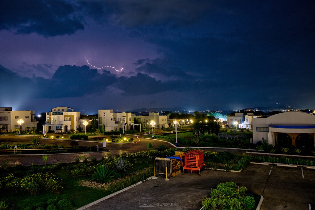 Lightning across a purple night sky above a quiet residential street with streetlights still lit