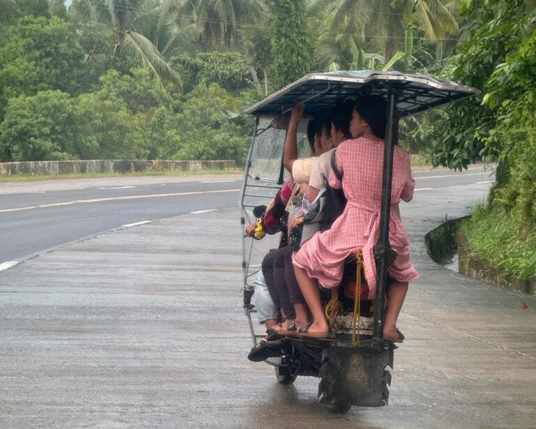A tricycle on a wet provincial road with the cab full and two passengers standing on the rear step, coconut palms on both sides