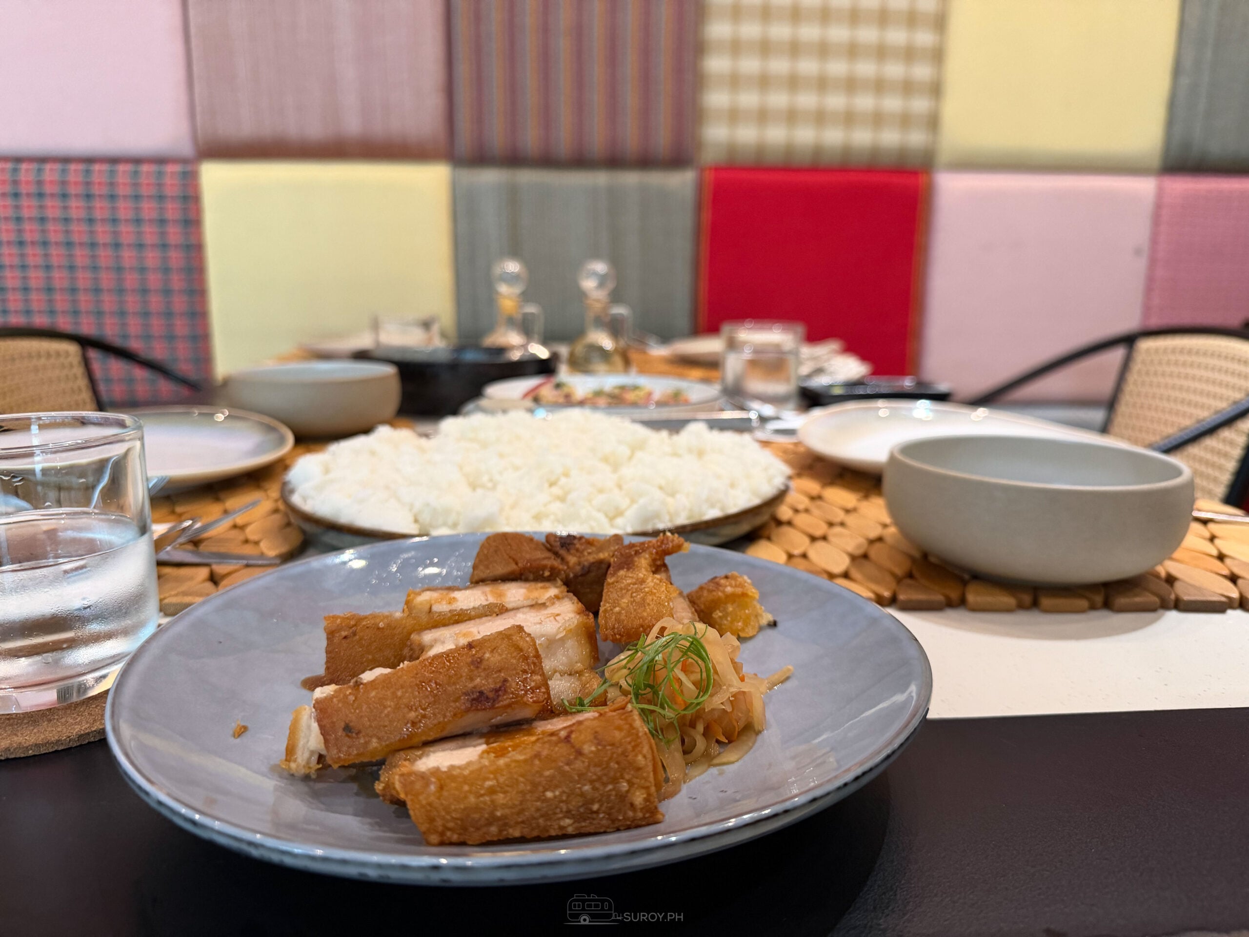 Slices of crispy pork belly on a gray plate at ATO-AH, with a bowl of rice and a patchwork fabric booth behind