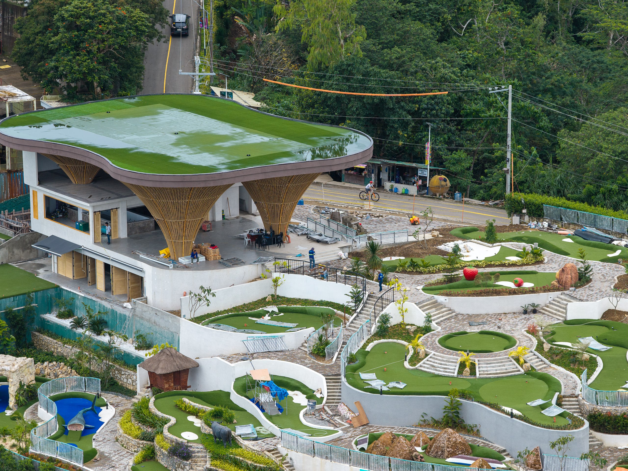 White building with a turf-covered roof on funnel-shaped columns, above terraced mini golf greens on a Busay hillside