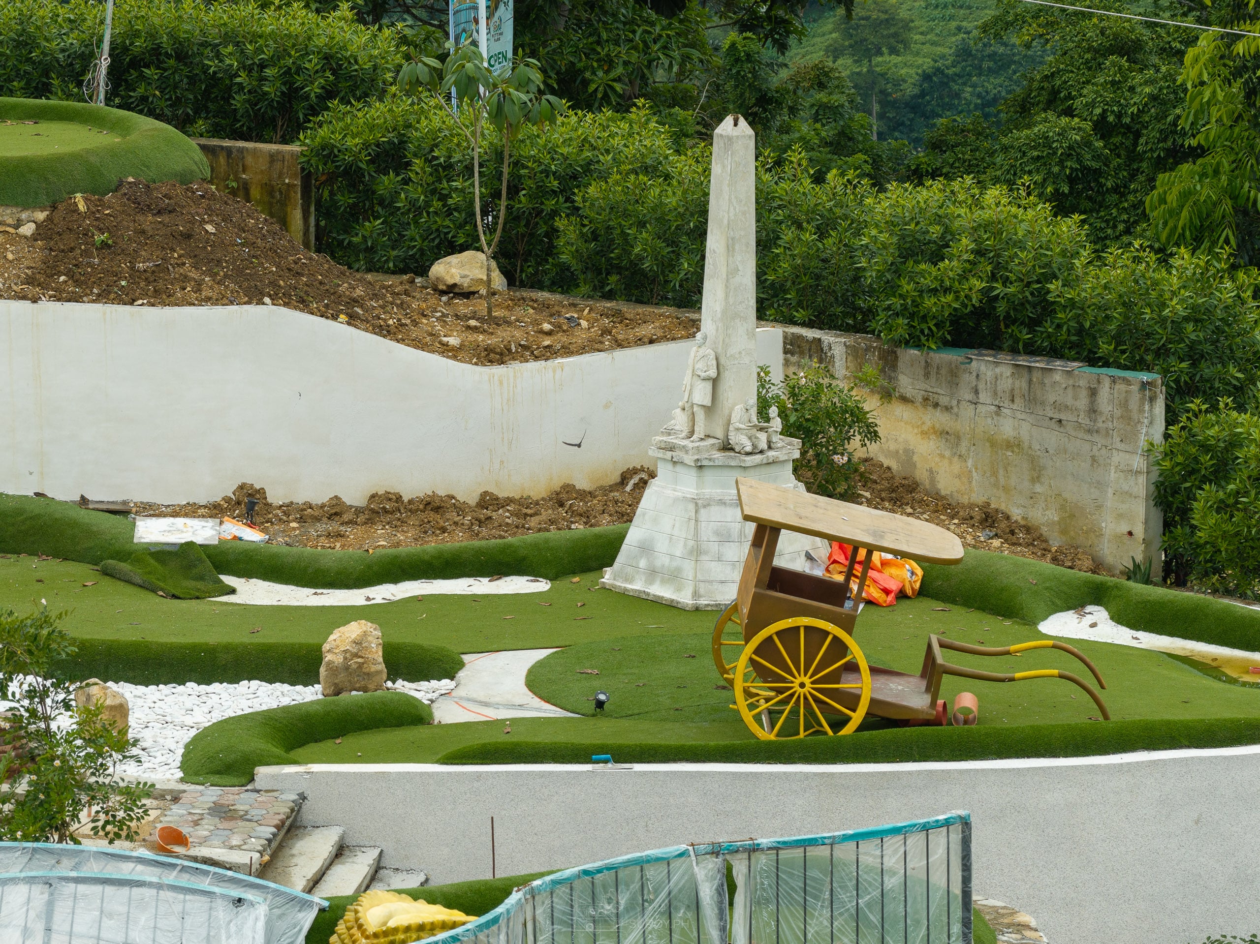 A yellow-wheeled kalesa beside a white obelisk on a mini golf green, with a bare dirt slope behind the wall