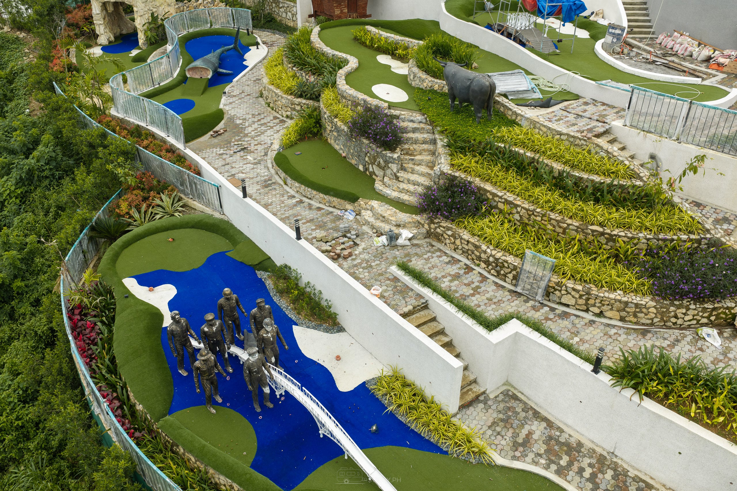 Terraced rows of plants stepping up a slope beside mini golf holes, with a group of dark statues wading through blue turf in the foreground