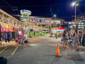 Lit Welcome Kalye Sugbo Market sign over a parking lot night market, with ukay clothing racks, green plastic tables, red food tents and the One Mango Avenue building behind