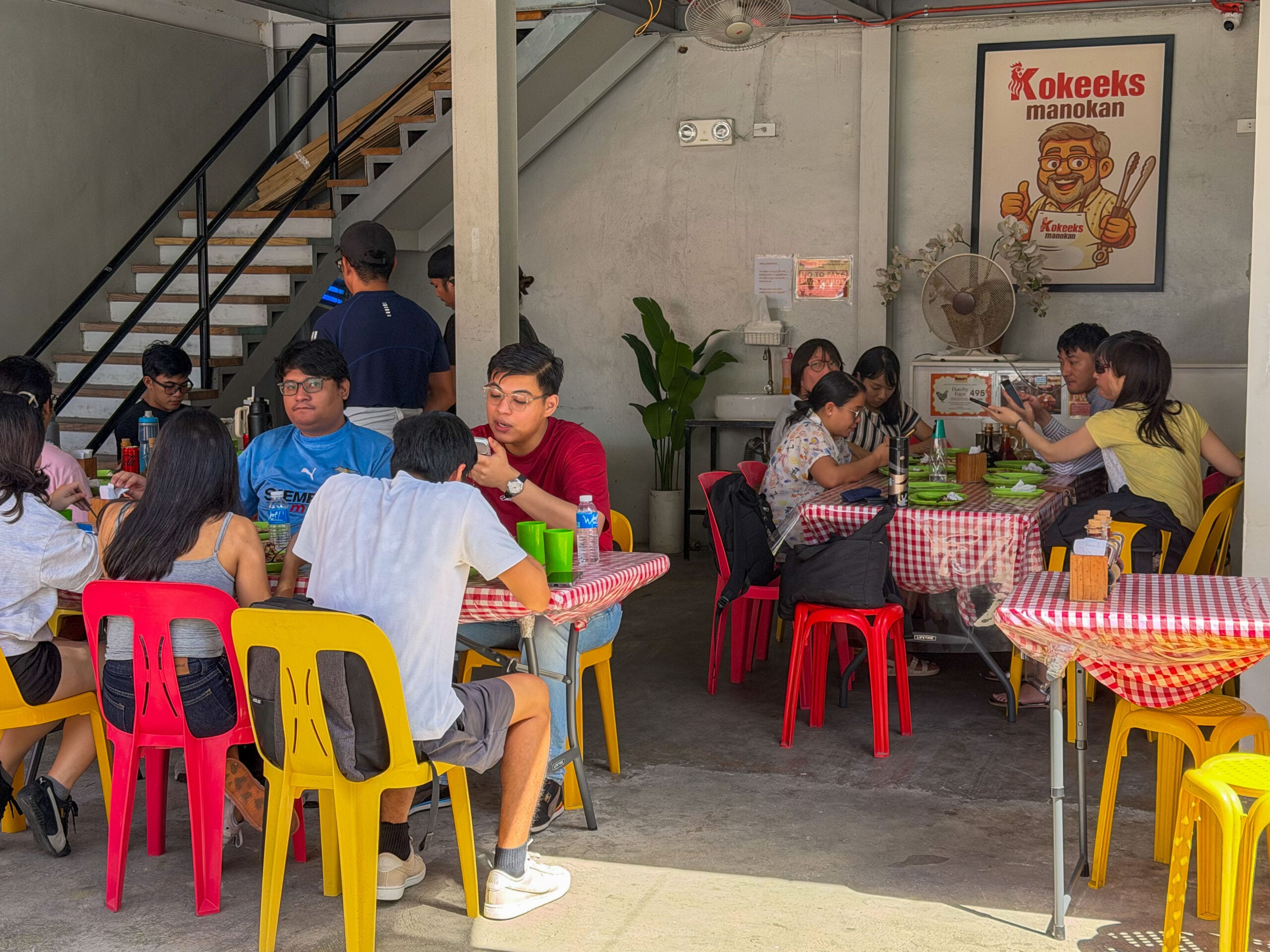 Full tables at Kokeek's Manokan with red and yellow plastic chairs, gingham tablecloths and a staircase to the upper level