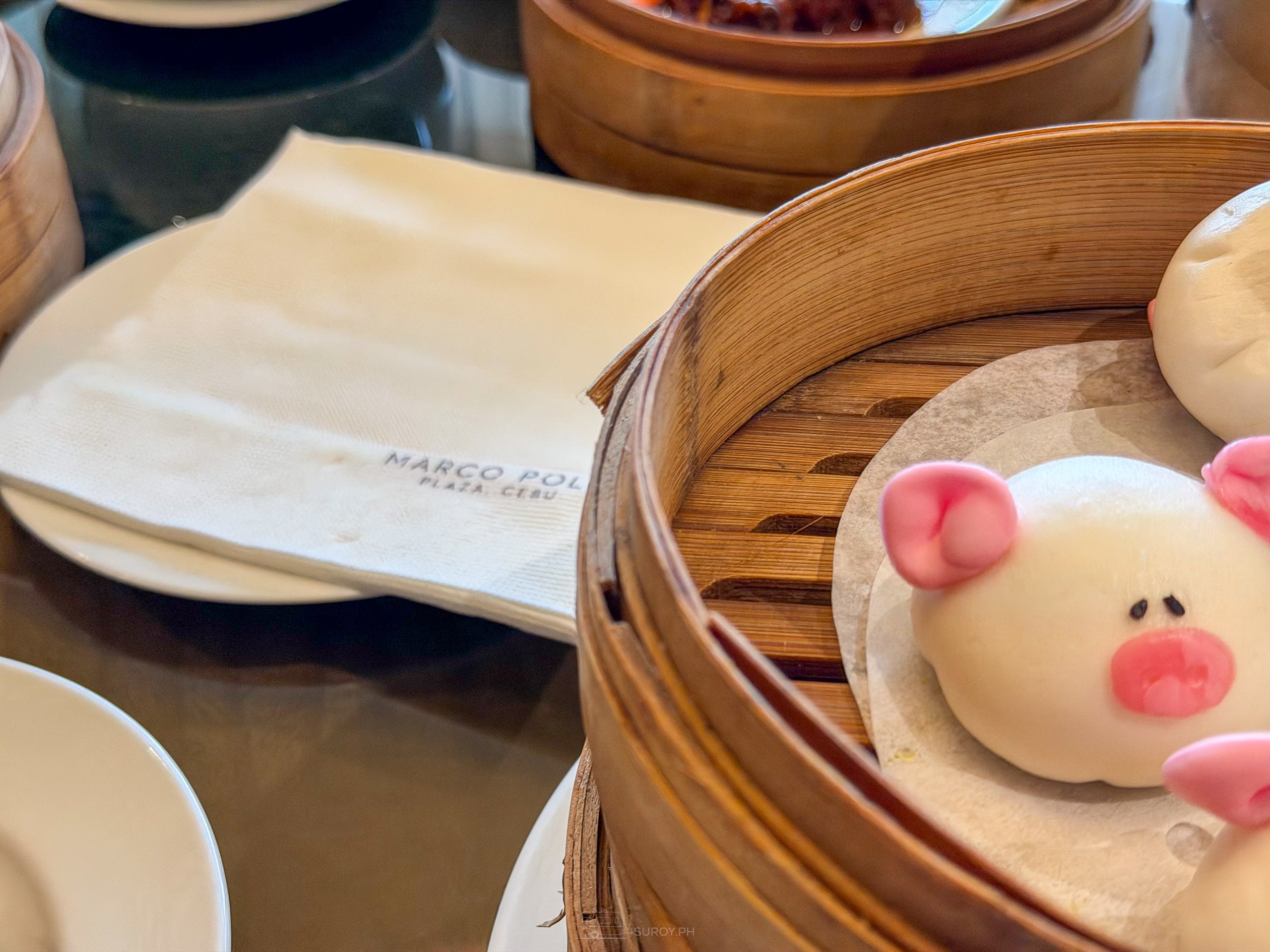 A white steamed bun shaped like a piglet, with pink ears and a pink snout, in a bamboo basket