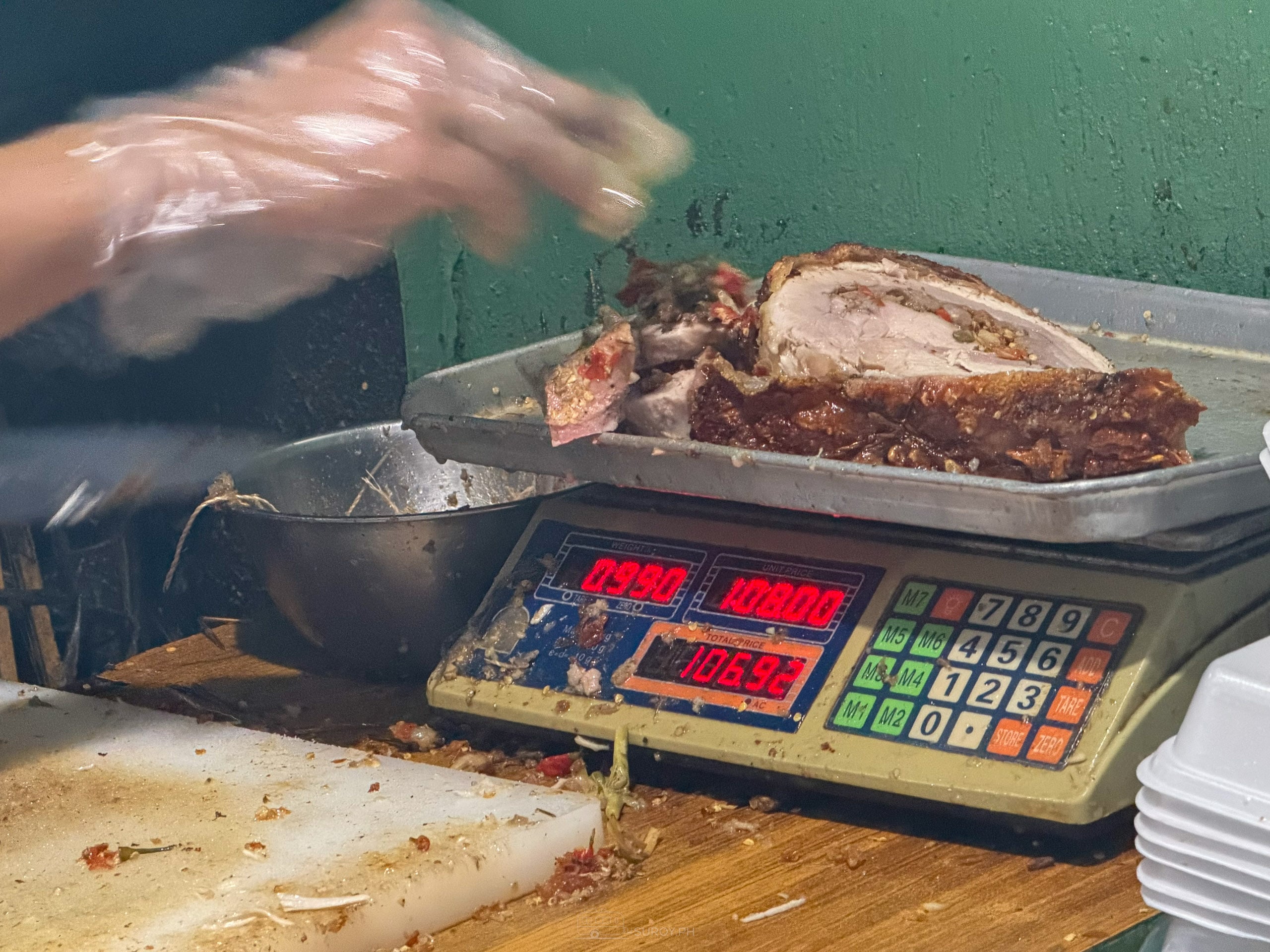 A slab of lechon belly on a digital scale reading 0.990 kilos at 1,080 pesos a kilo, total 1,069.20 pesos